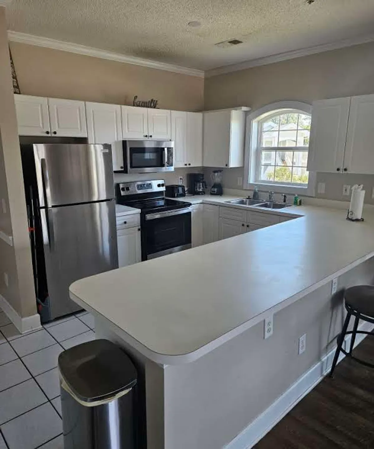 This image features a warm, welcoming kitchen with white cabinetry, a spacious island countertop, a sink overlooking a window with treetop views, and modern stainless steel appliances. It evokes the feeling of family meal prep and social gatherings, with a layout ideal for both cooking and entertaining.