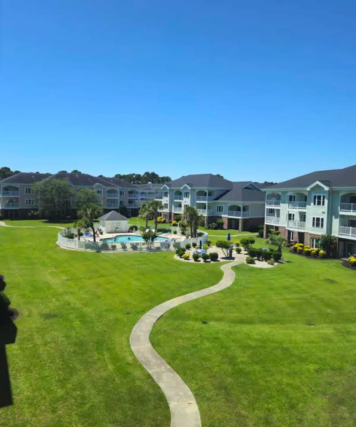 The hero image showcases the condo complex exterior, featuring a sparkling swimming pool surrounded by sun loungers, manicured green lawns, palm trees, and walking paths. In the background, attractive low-rise condo buildings add to the inviting, resort-like feel. The scene is vibrant and sunlit, suggesting a lively yet relaxing atmosphere ideal for vacationers.