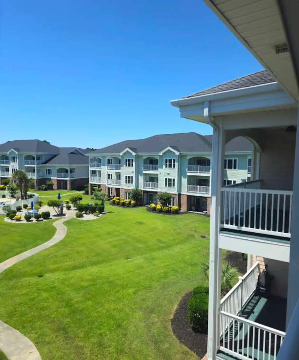A wider exterior view of the apartment building, showcasing multiple balconies overlooking manicured green lawns and paved walking paths. The landscaping and distant sunset lighting evoke a relaxed, scenic atmosphere, highlighting the private outdoor spaces available to residents.