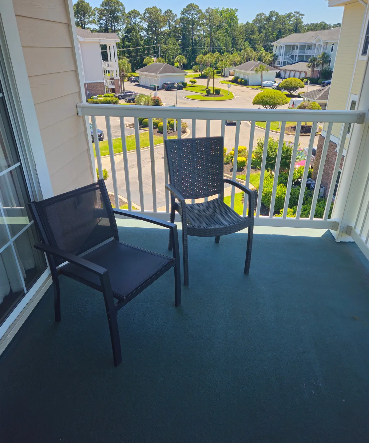 A tranquil private balcony with two comfortable chairs overlooks lush green trees and a peaceful neighborhood. Soft sunlight bathes the space, suggesting the perfect spot for morning coffee or evening relaxation. The view highlights treetop vistas and the surrounding community, enhancing the sense of privacy and serenity.