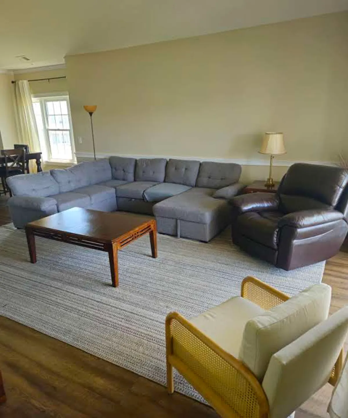A comfortable living area featuring a sectional sofa, coffee table, armchair, and a window with curtains. The setup is arranged for casual gatherings, movie nights, or quiet reading sessions, reinforcing the feeling of a communal haven.