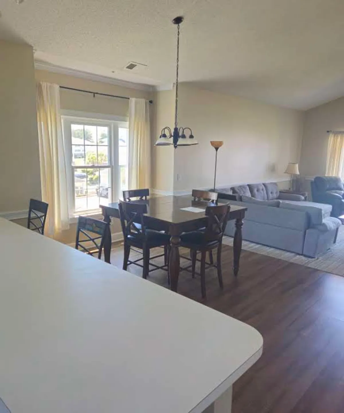 A spacious open-plan area combining a comfortable sofa, a dining table, and large windows that allow abundant natural light and offer a view of the outdoors. The soft furnishings and layout encourage both relaxation and social interaction.