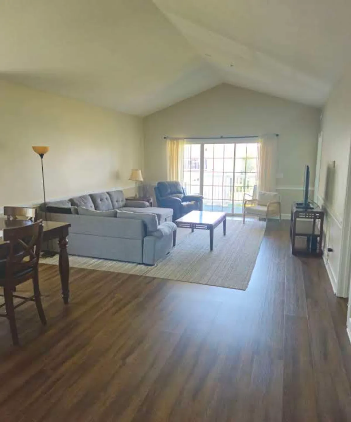 A cozy living room scene showing a sofa, coffee table, window, and a dining table in the background. Sunlight pours in through the window, creating a warm and inviting atmosphere ideal for both relaxation and socializing.
