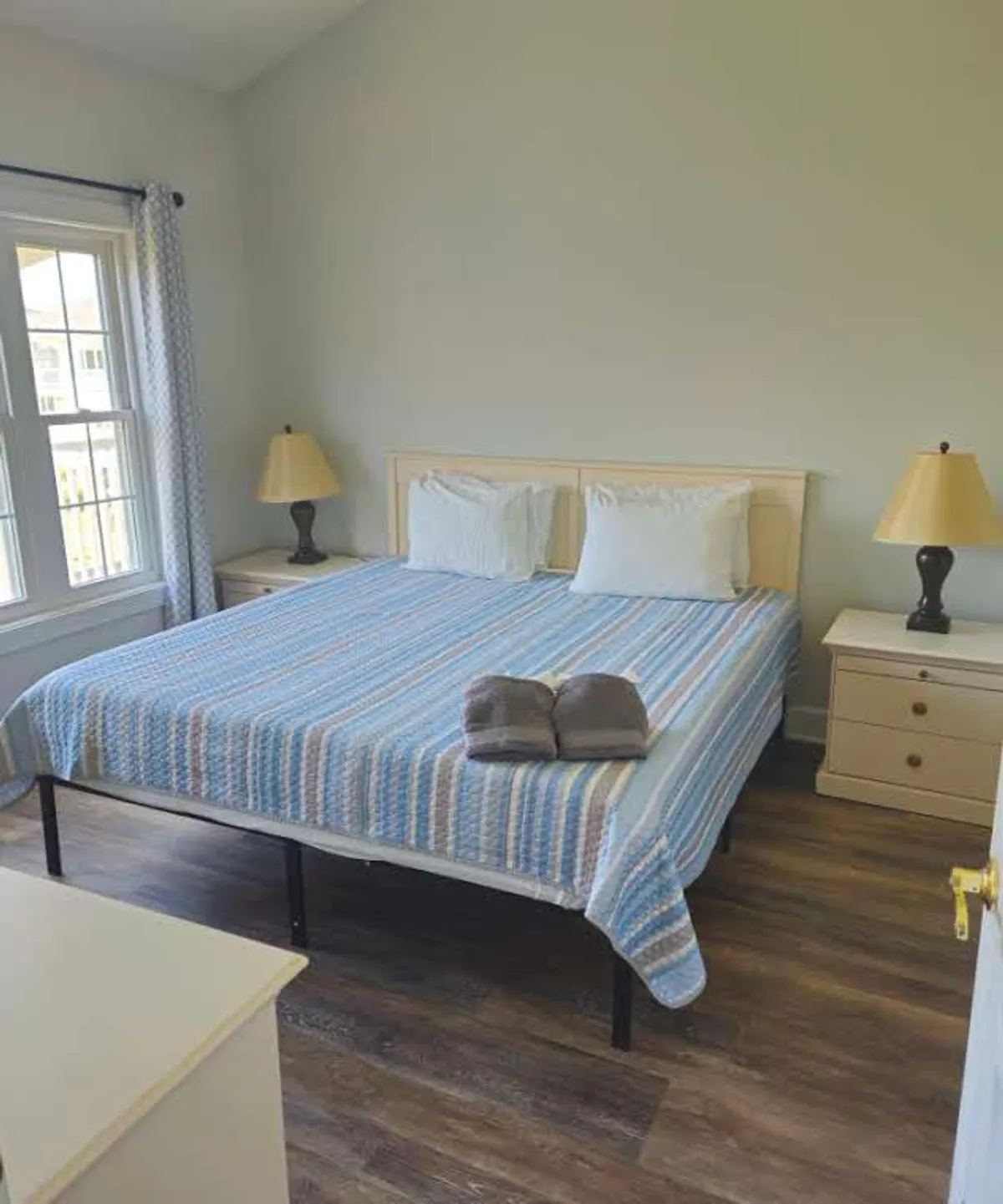 A bright, comfortable bedroom featuring a bed with a blue striped blanket, flanked by nightstands with lamps. Sunlight streams in through a large window, creating a serene and inviting atmosphere ideal for rest and relaxation.
